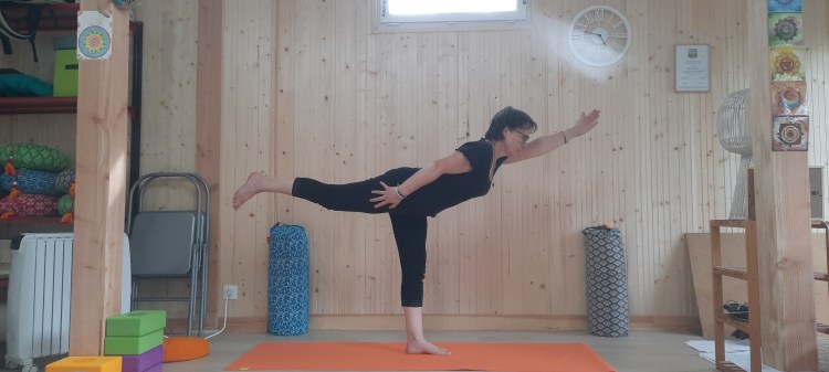Une femme pratiquant une posture de yoga sur un tapis orange dans une salle en bois, avec des accessoires de yoga colorés en arrière-plan.