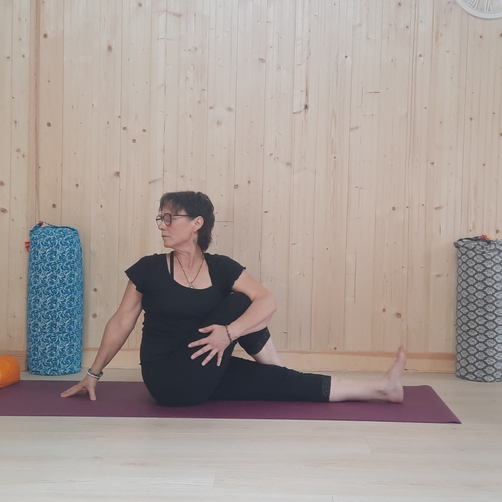 Femme pratiquant une posture de yoga assise sur un tapis dans une salle au mur en bois, avec des accessoires de yoga à proximité.